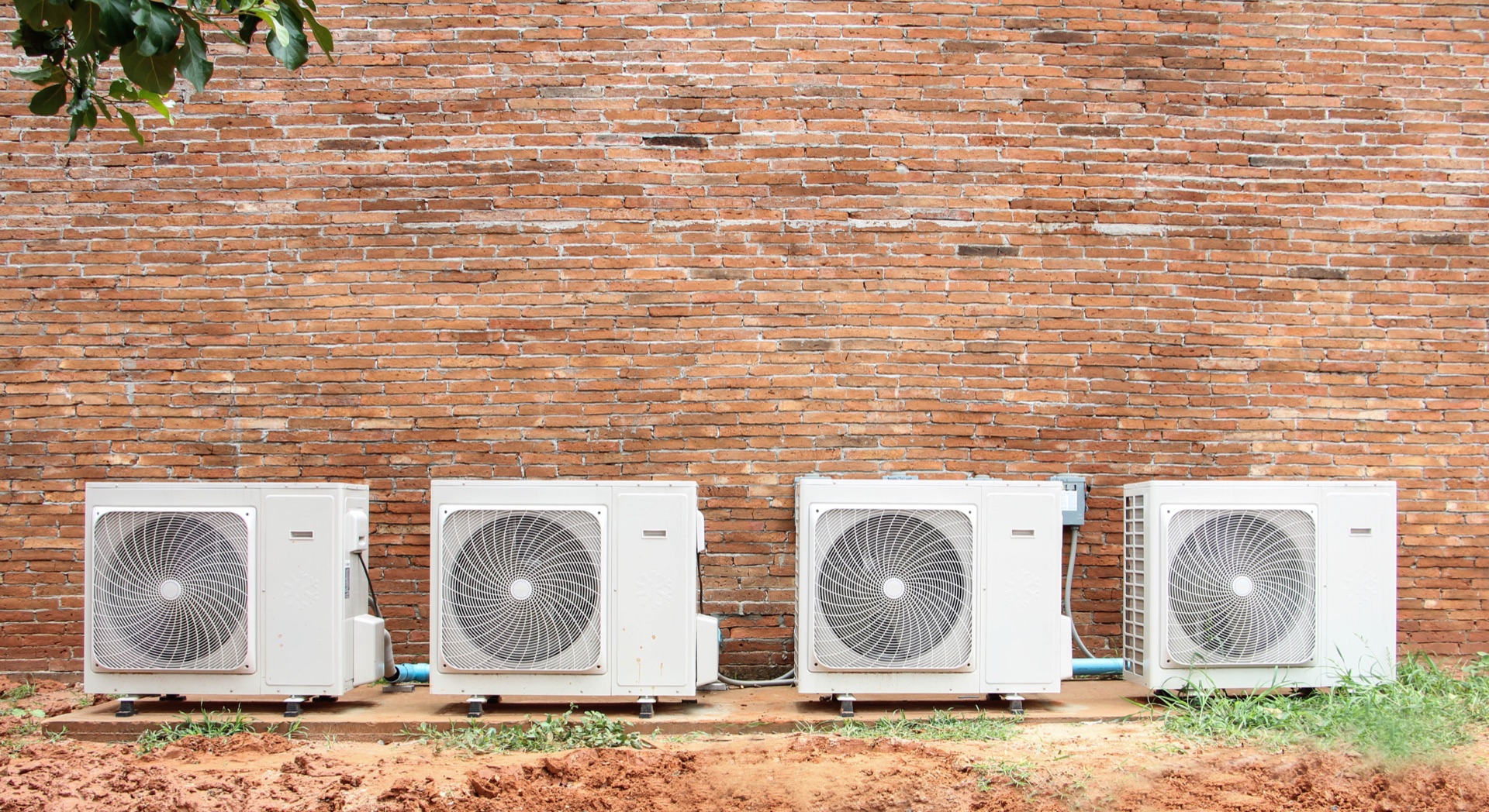 Four white air conditioning units mounted against a brick wall with dirt ground below