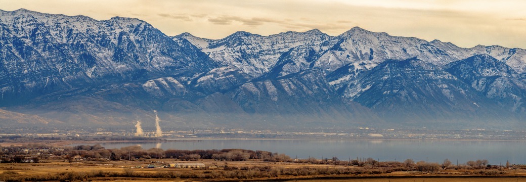 Snow-capped mountains rising behind a wide lake with industrial smokestacks visible in the valley below
